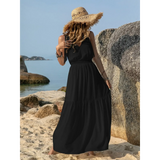 Woman wearing a black maxi dress and straw hat standing on a beach with rocks in the background