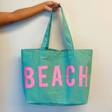 Teal Canvas Beach Tote bag with pink BEACH lettering and long handles held by hand