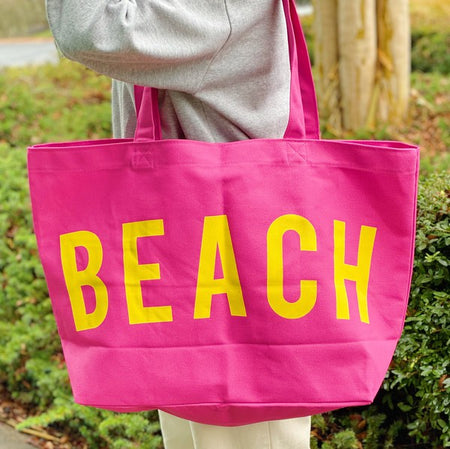 Bright pink Canvas Beach Tote with yellow BEACH text, carried on shoulder outdoors