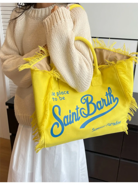 Yellow large resort tote bag with fringe and blue Saint Barth text carried by person in beige sweater