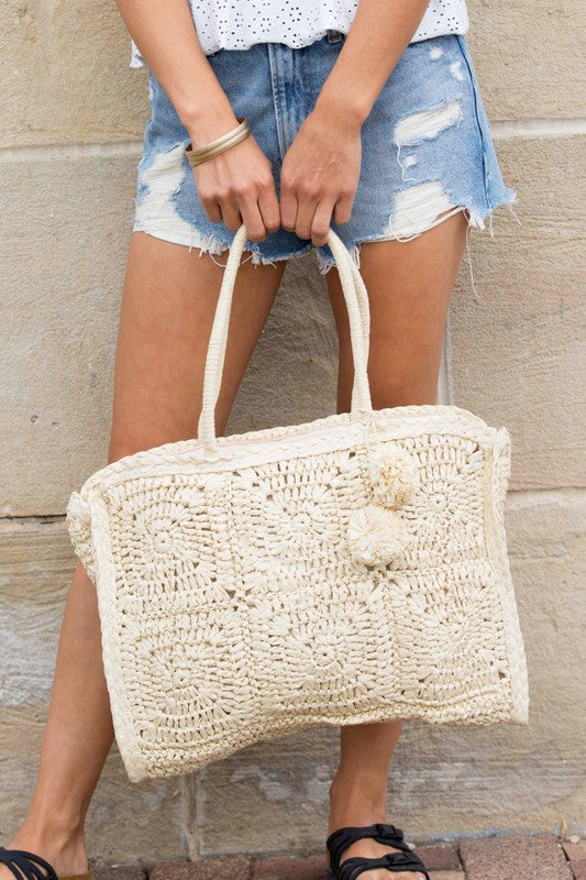 Babe Beachwear Straw Summer Tote bag in natural woven straw with pom pom details held by woman in denim shorts