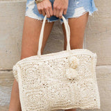 Babe Beachwear Straw Summer Tote bag in natural woven straw with pom pom details held by woman in denim shorts