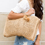 Babe Beachwear straw summer tote bag with pom pom details carried by woman in white top and denim shorts