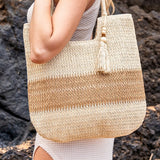 Two tone straw tote bag by Babe Beachwear with beige and natural brown stripes held by woman in white dress