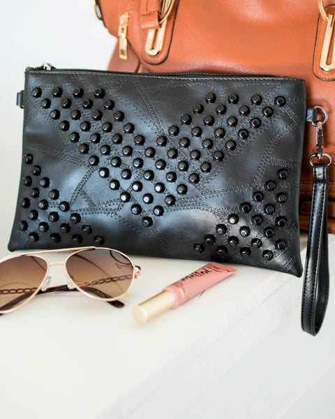 Black vacation evening clutch for women with studded design and wrist strap displayed with sunglasses and lip gloss