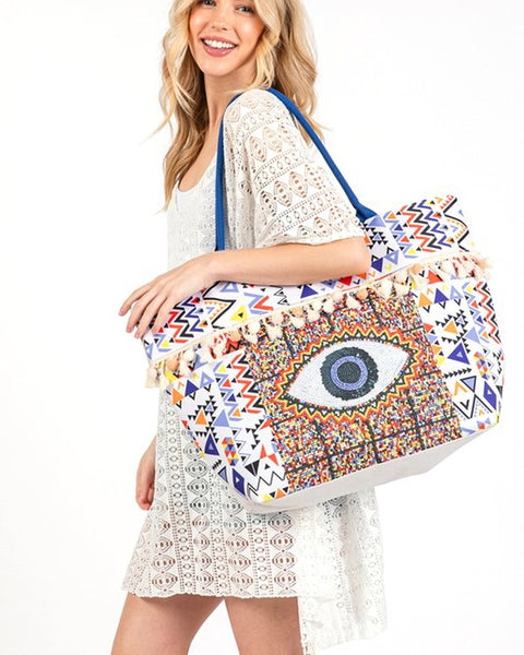 Babe Beachwear Pom Pom Evil Eye bag with colorful geometric patterns and blue straps worn by woman in white dress