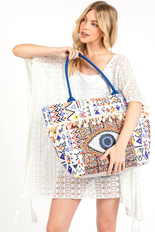 Pom Pom Evil Eye bag by Babe Beachwear with colorful geometric patterns and blue handles worn by a woman in a white lace cover-up