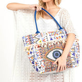 Pom Pom Evil Eye bag by Babe Beachwear with colorful geometric patterns and blue handles worn by a woman in a white lace cover-up