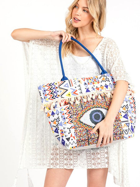Pom Pom Evil Eye bag by Babe Beachwear with colorful geometric patterns and blue handles worn by a woman in a white lace cover-up