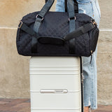 Zahra carry on overnight bag in black by Babe Beachwear placed on white rolling suitcase