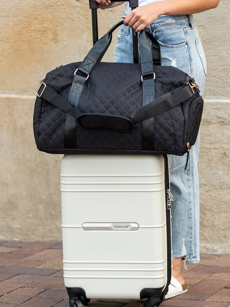 Zahra carry on overnight bag in black by Babe Beachwear placed on white rolling suitcase